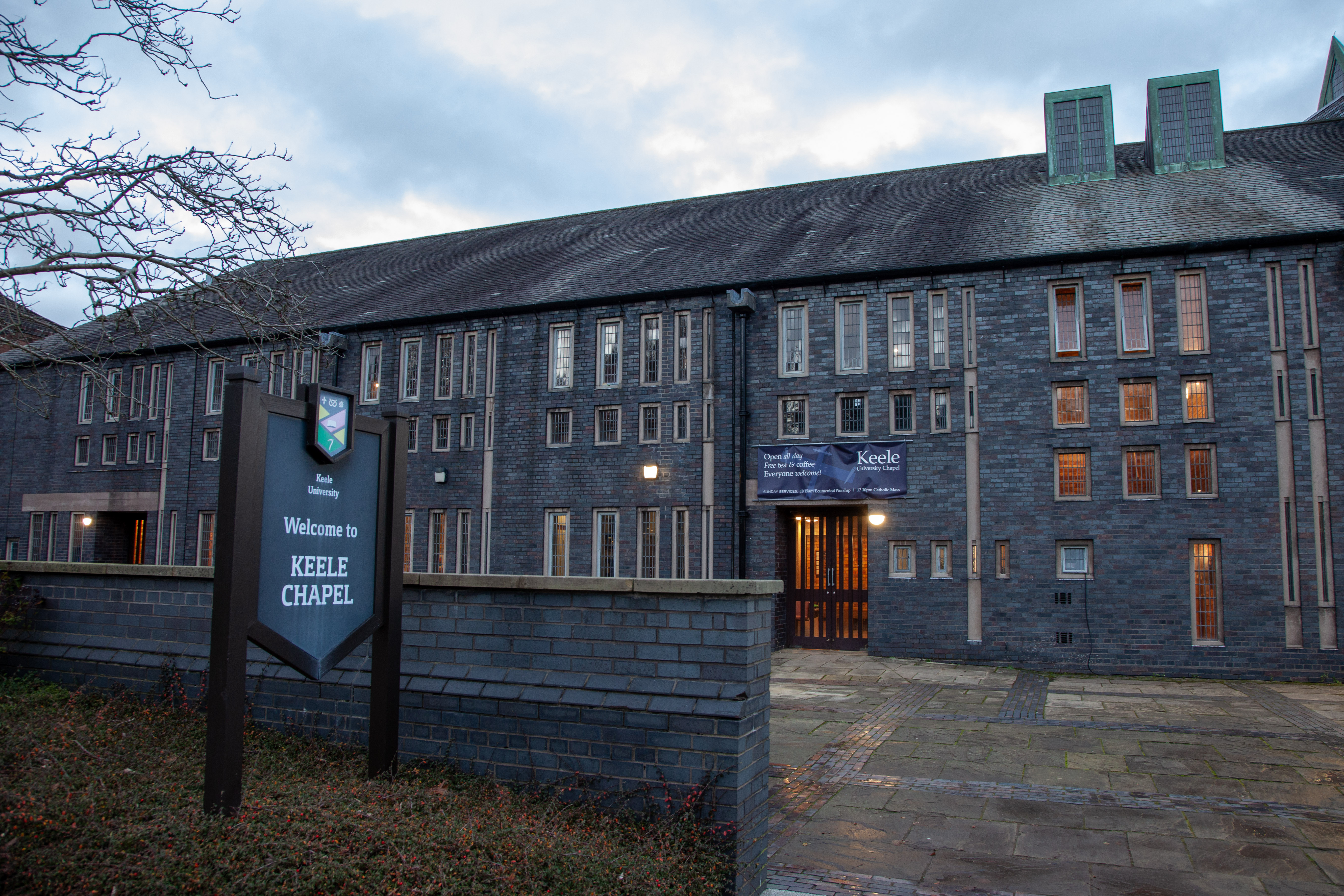Image of Keele University Chapel. A tall, dark grey brick building with many long vertical windows and a slate root